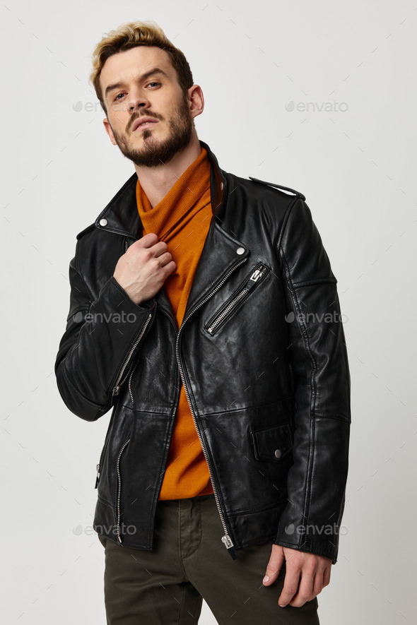 attractive young man in an orange sweater and in a leather jacket on a light background touches the