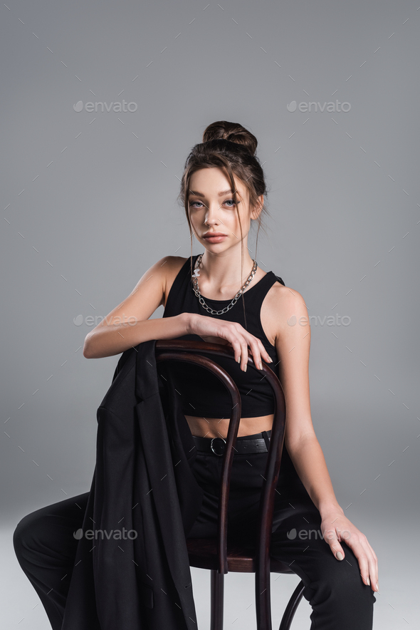 young woman in black crop top and silver necklace sitting on chair near jacket isolated on grey
