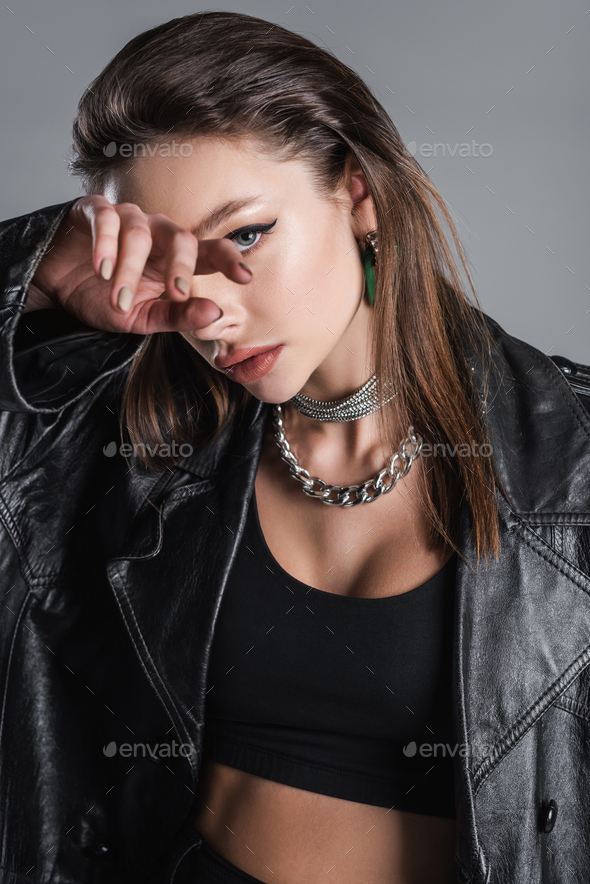 young woman in black leather coat and silver necklaces obscuring face with hand isolated on grey
