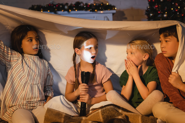 Girl holding flashlight near scared interracial friends under blanket and christmas decor at home