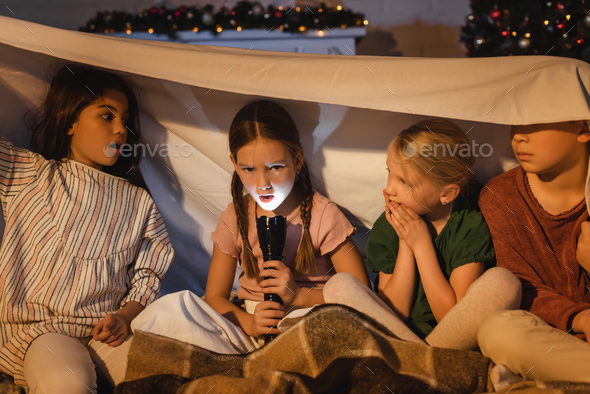 Girl holding flashlight near scared friends under blanket during christmas at home