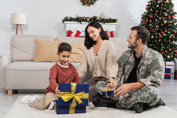 happy boy opening gift box while sitting on floor near smiling mother and father in military uniform