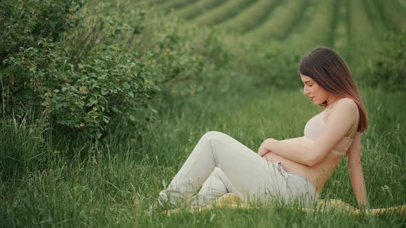 Pregnant young woman with dark hair in pink top and light pants sitting on green grass in field