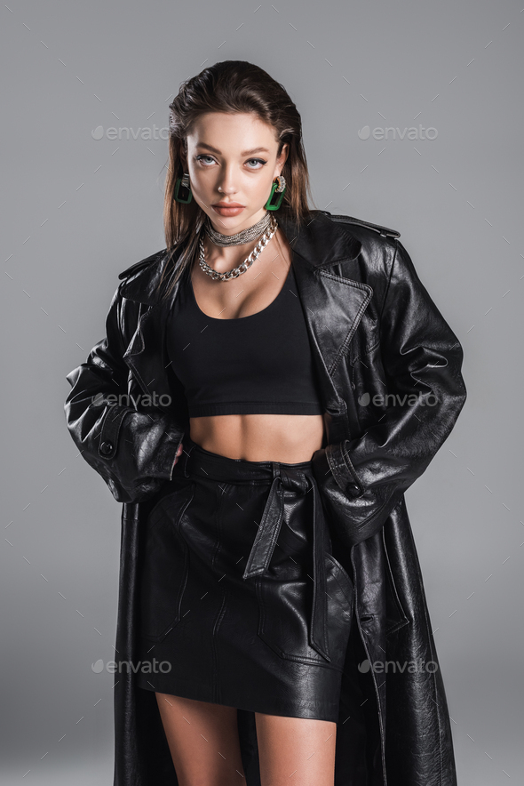 brunette woman in black crop top and leather jacket standing with hands in pockets isolated on grey