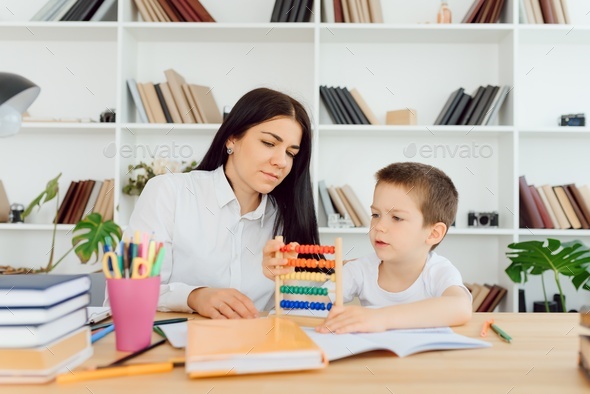 Female private tutor helping young student with homework at desk in bright child\'s room