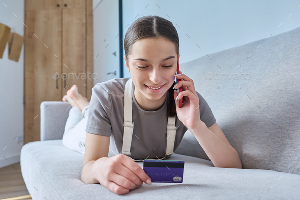 Teenage girl with credit bank card talking on cell phone lying on sofa at home