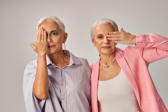 trendy mature models in blue and pink wear obscuring faces with hands and looking at camera on grey