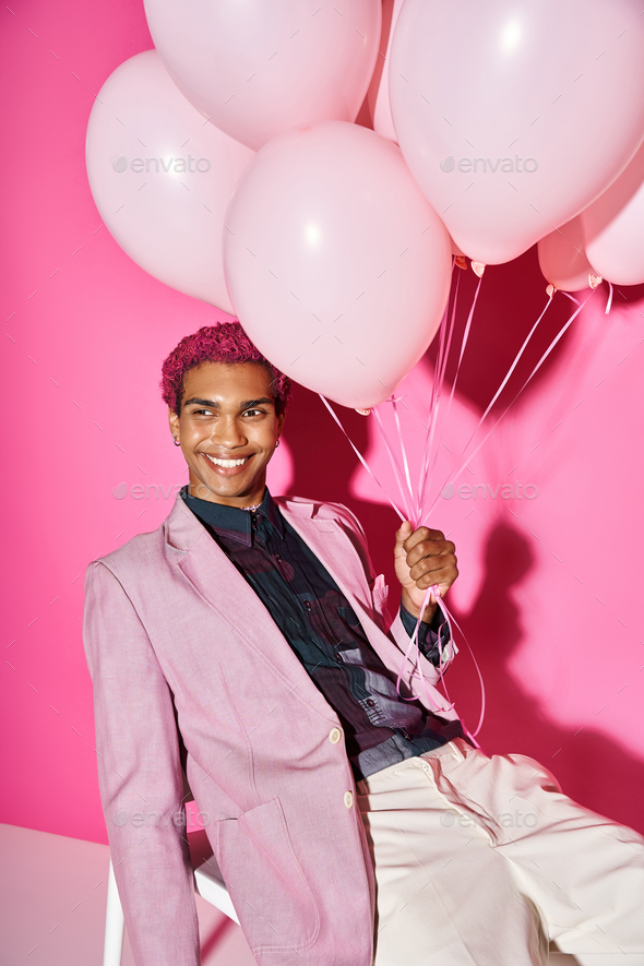 good looking man with curly pink hair posing on white chair with balloons in hand, doll like