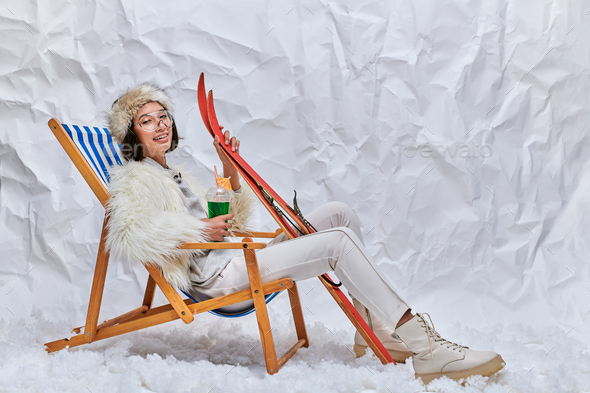 happy asian model in winter clothes sitting in deck chair with cocktail and skis in snowy studio