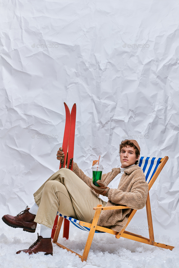 trendy man in warm outfit sitting in deck chair with hot toddy cocktail and drink in snowy studio