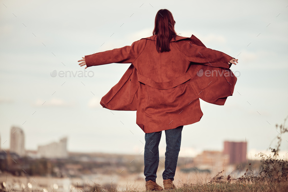 Long-haired girl in red coat fluttering in wind stands with back to camera, arms outstretched