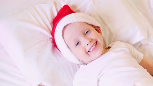 Child wakes up before Christmas. Little boy in Santa hat and pajamas lying in white bed