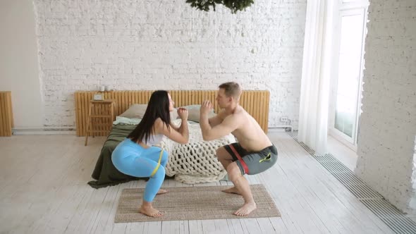 Young Sporty Man and Woman Are Doing Squats Together Using Rubber Bands at Home alt