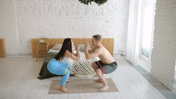 Young Sporty Man and Woman Are Doing Squats Together Using Rubber Bands at Home