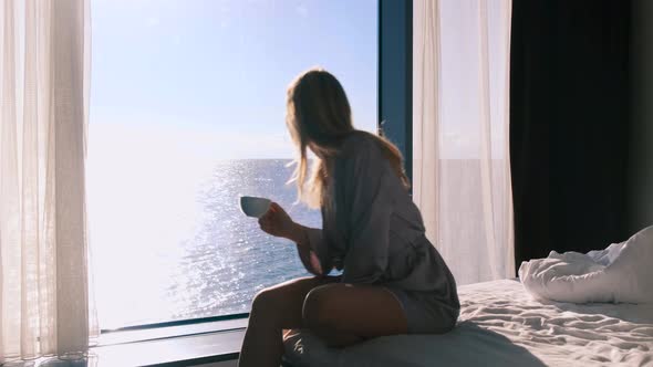 Peaceful Happy Young Blonde Woman in Bathrobe Enjoys Her Morning Coffee Sitting on Bed Against