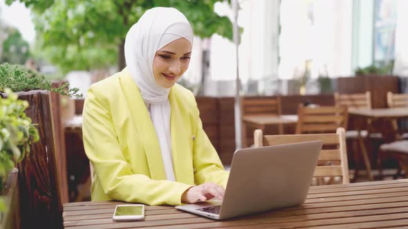 Smiling Young Muslim Woman Wearing Headscarf Sits in Cafe and Uses Laptop