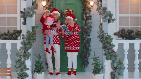 Senior Grandmother Grandfather with Granddaughter Standing at Christmas House Porch Waving Hello Hi alt