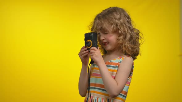 Cute Girl is Using Retro Photo Camera for Shooting Pictures in Yellow Studio alt