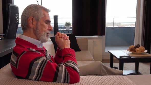 An Elderly Man Sits on a Couch in an Apartment Living Room, Watches TV and Acts Upset alt