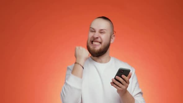 Happy Young Bearded Man Is Looking on Smartphone Doing Winner Gesture