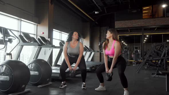 Two Lovely Plus Size Female Athletes Squatting with Kettlebells at the Gym