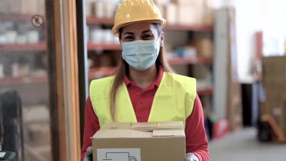 Mature woman working inside warehouse holding delivery package while wearing protective face mask
