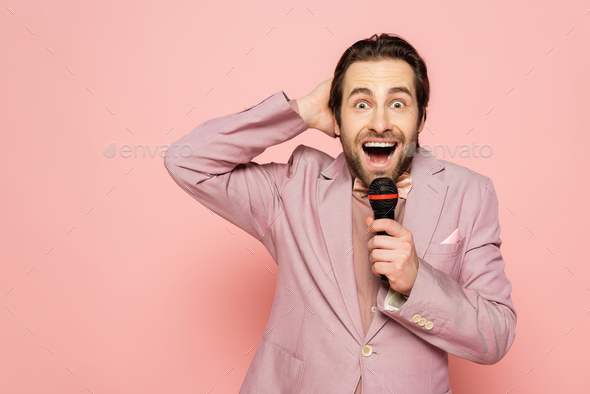 Excited host of event in jacket holding microphone and hand near head isolated on pink
