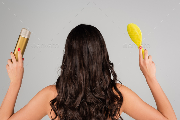 back view of curly woman holding hair spray and yellow hair brush isolated on grey
