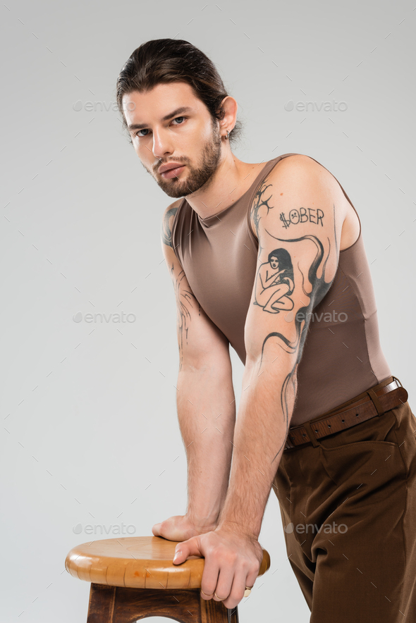 Long haired tattooed man standing near chair isolated on grey