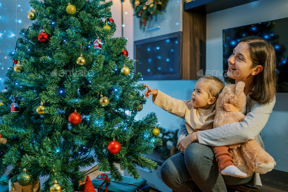 Little smiling girl pointing her finger at a sparkling Christmas tree while sitting on her mother