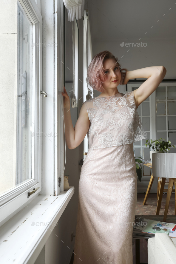 Romantic lady, red hair woman in white vintage dress stands next to window.