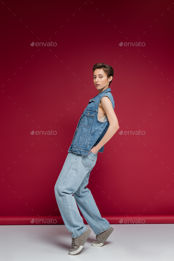 full length of stylish model in denim outfit with vest posing with hands in back pockets on jeans on