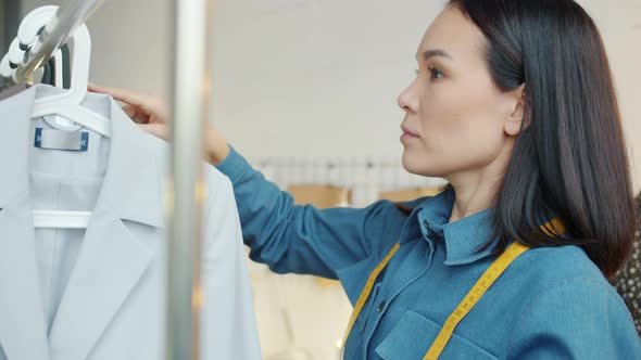 Asian Lady Fashion Designer Hanging New Clothes on Rails in Dressmaking Studio alt