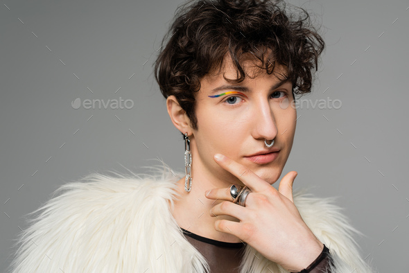 portrait of brunette nonbinary person with rainbow eye liner and silver accessories holding hand