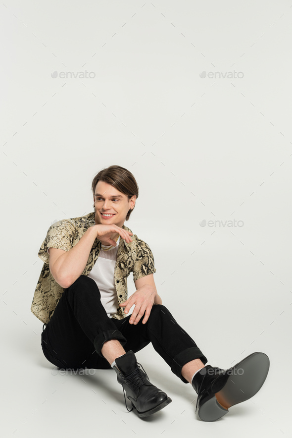 full length of carefree bigender person in black trousers and snakeskin print blouse sitting on grey