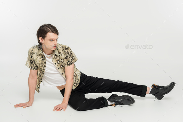 full length of stylish pansexual person in snakeskin print blouse and black pants posing on grey
