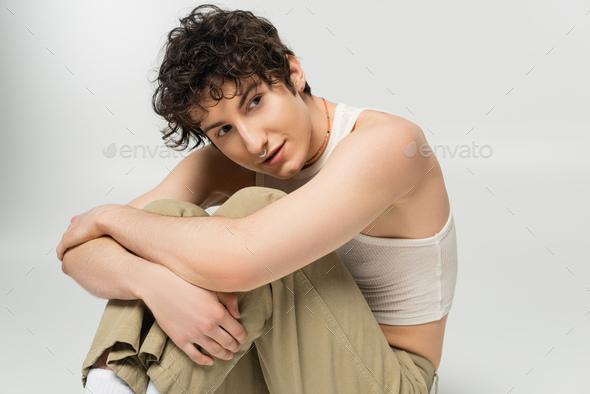 young pansexual person with wavy brunette hair hugging knees while sitting and looking away isolated