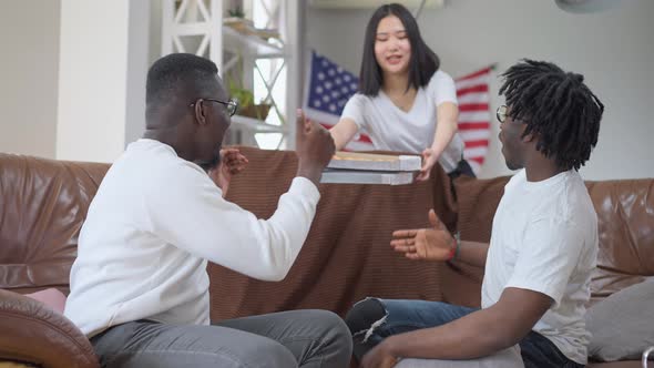 African American Men Talking Sitting on Couch As Asian Woman Bringing Delivered Pizza alt