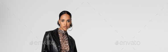 stylish african american woman in trendy leather jacket and hoop earrings isolated on grey, banner