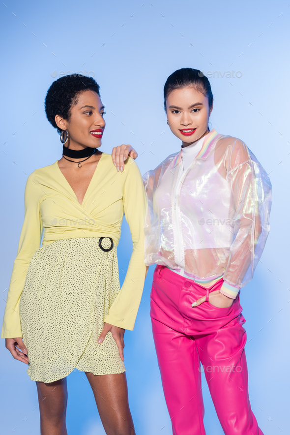 Carefree asian woman with red lips posing with african american friend on blue background