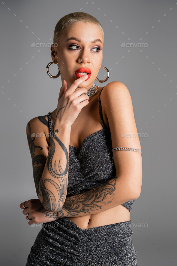 seductive tattooed woman in hoop earrings and lurex crop top touching red lips and looking away
