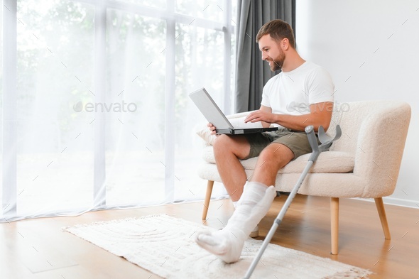 Happy young man with arm in a cast sitting on the couch at home and communicating on a laptop