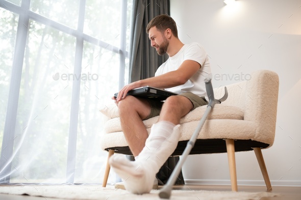 Happy young man with arm in a cast sitting on the couch at home and communicating on a laptop