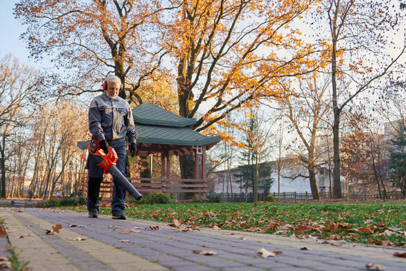 Gray haired maintenance worker cleaning sidewalk with leaf blower.