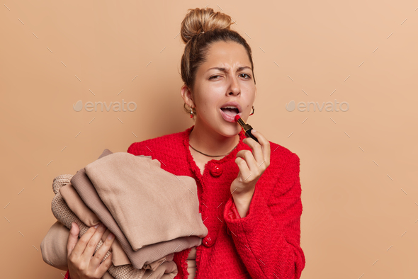 Displeased woman with fair hair gathered in bun being busy folding washed clothes applies lipstick