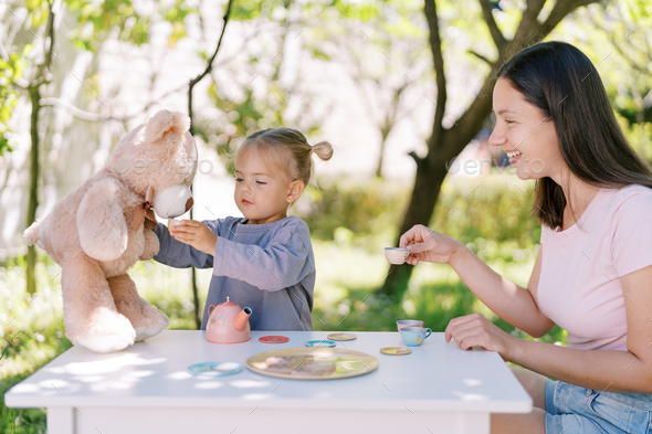 Smiling mom with toy cup in hand watches little girl giving tea to teddy bear at table in garden