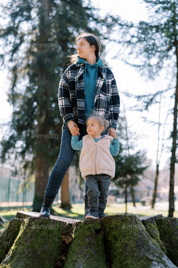 Mom with a little girl stand on a huge stump in a sunny meadow and look away