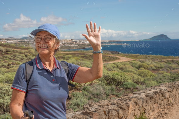 Cheerful senior woman with backpack outdoors on hike in countryside with sea view
