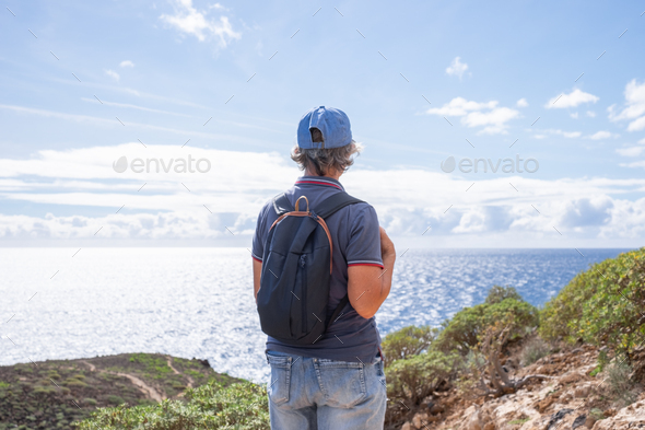 Back view of senior woman with backpack outdoors on hike in countryside with sea view