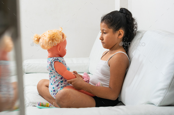 sad brown girl, sitting next to her doll on the living room sofa, looking at the doll from the front